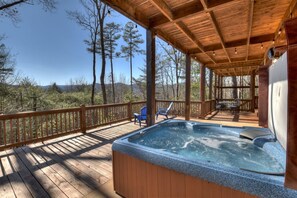 Une terrasse en bois avec un jacuzzi, des chaises et une table, le tout installé sous une zone abritée offrant une vue sur les arbres et les montagnes.