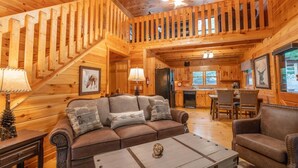 Een houten chalet interieur met een trap, een woonkamer met bank en salontafel, een keukenhoek, en een eettafel met stoelen.