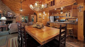 L’intérieur d’un chalet en bois comprend une table à manger, des chaises, une cheminée, un coin cuisine et un lustre.