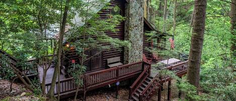 A rustic cabin with a stone chimney, wooden deck, and a staircase leading to the entrance.