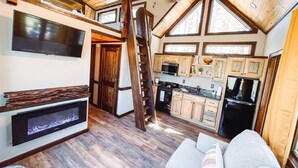 A cozy cabin interior with a loft bedroom, a fireplace, a kitchen with wooden cabinets, and a large window. - Misty Mountain Treehouse- Blue Ridge GA (Morganton)