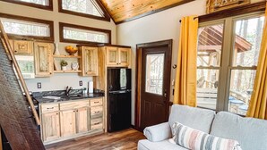 A cozy kitchen with wooden cabinets, a black refrigerator, and a dining area with a gray sofa and yellow curtains. - Misty Mountain Treehouse- Blue Ridge GA (Morganton)