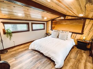 Une chambre avec un plafond en bois, un grand lit, deux fenêtres et une table de chevet avec une lampe.