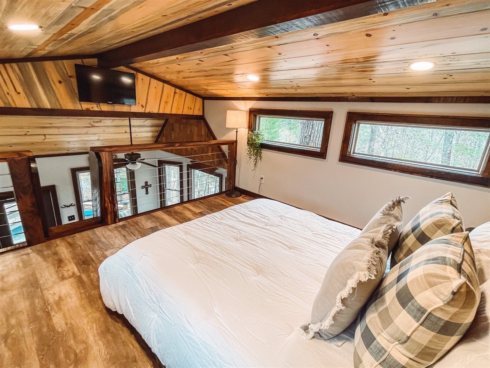 Un dormitorio con techo de madera, una cama con ropa blanca y dos ventanas con vistas a árboles.