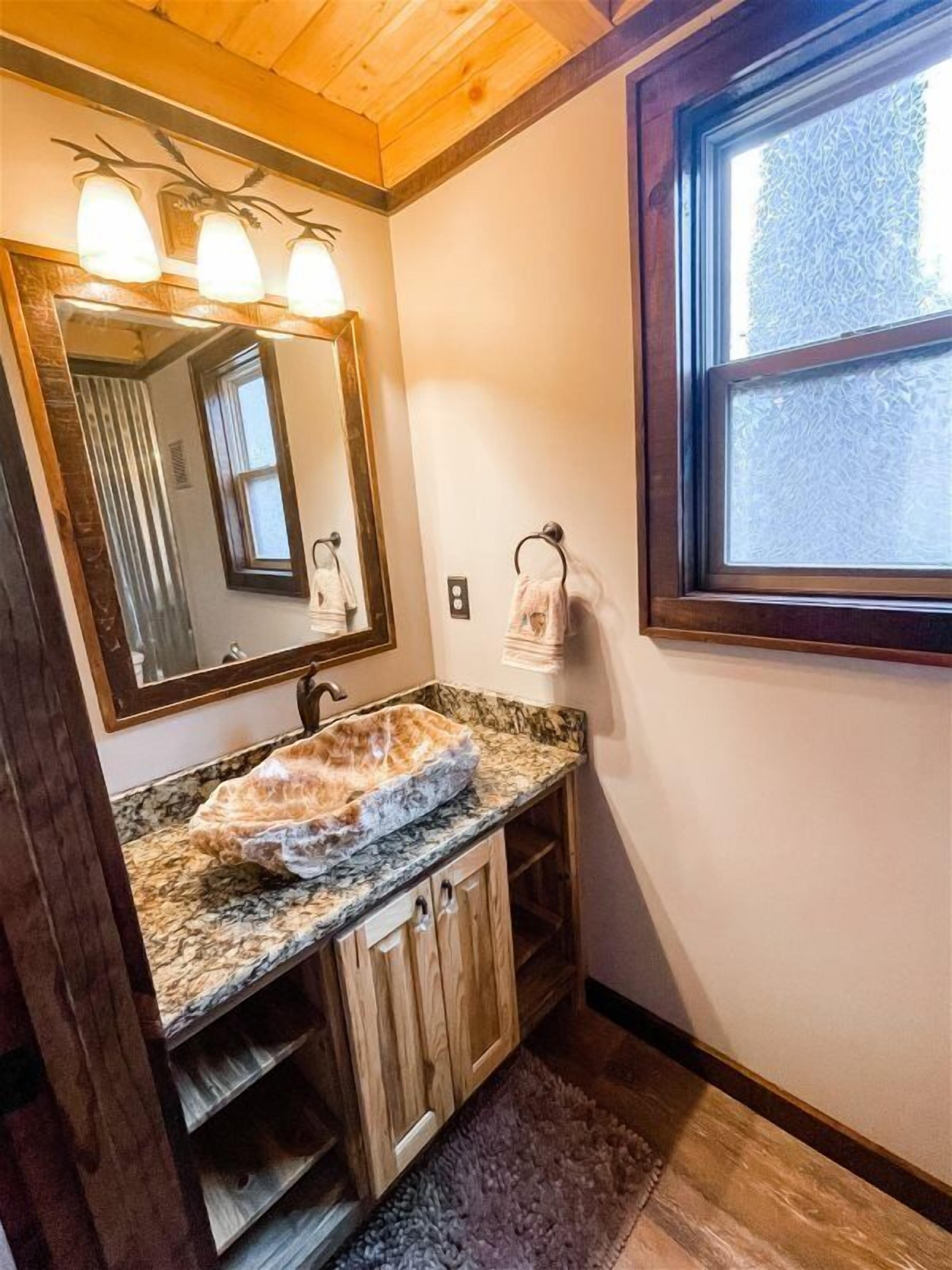 Un baño con lavamanos de encimera de granito, armarios de madera, un espejo grande y una ventana con vidrio esmerilado.