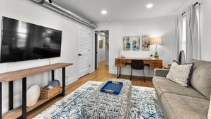 Una sala de estar moderna con una televisión de pantalla plana, una mesa de centro de madera, un tapete estampado, un sofá y un escritorio con una lámpara.