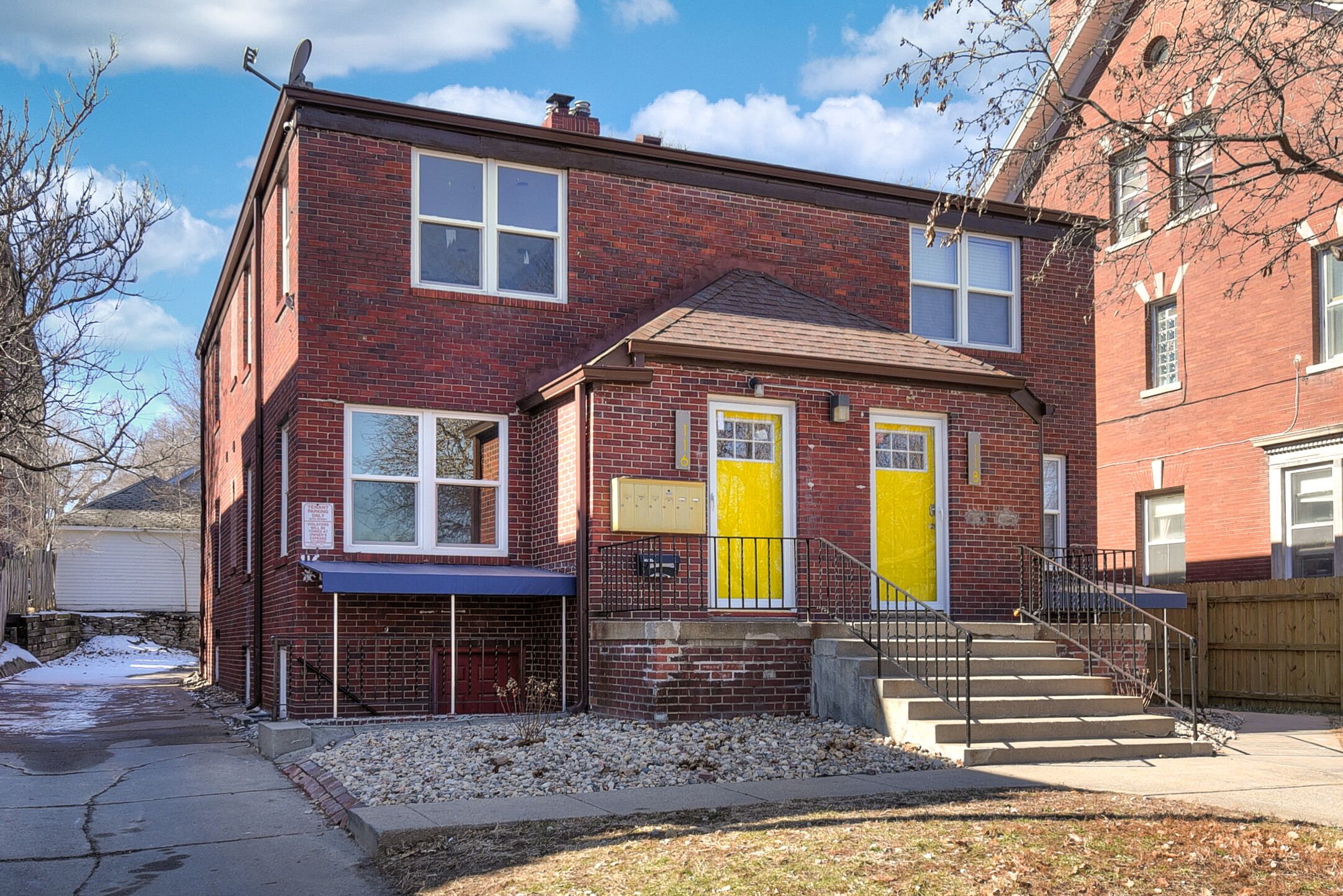 A two-story brick house with a blue awning, a yellow door, and a set of stairs leading to the front door.