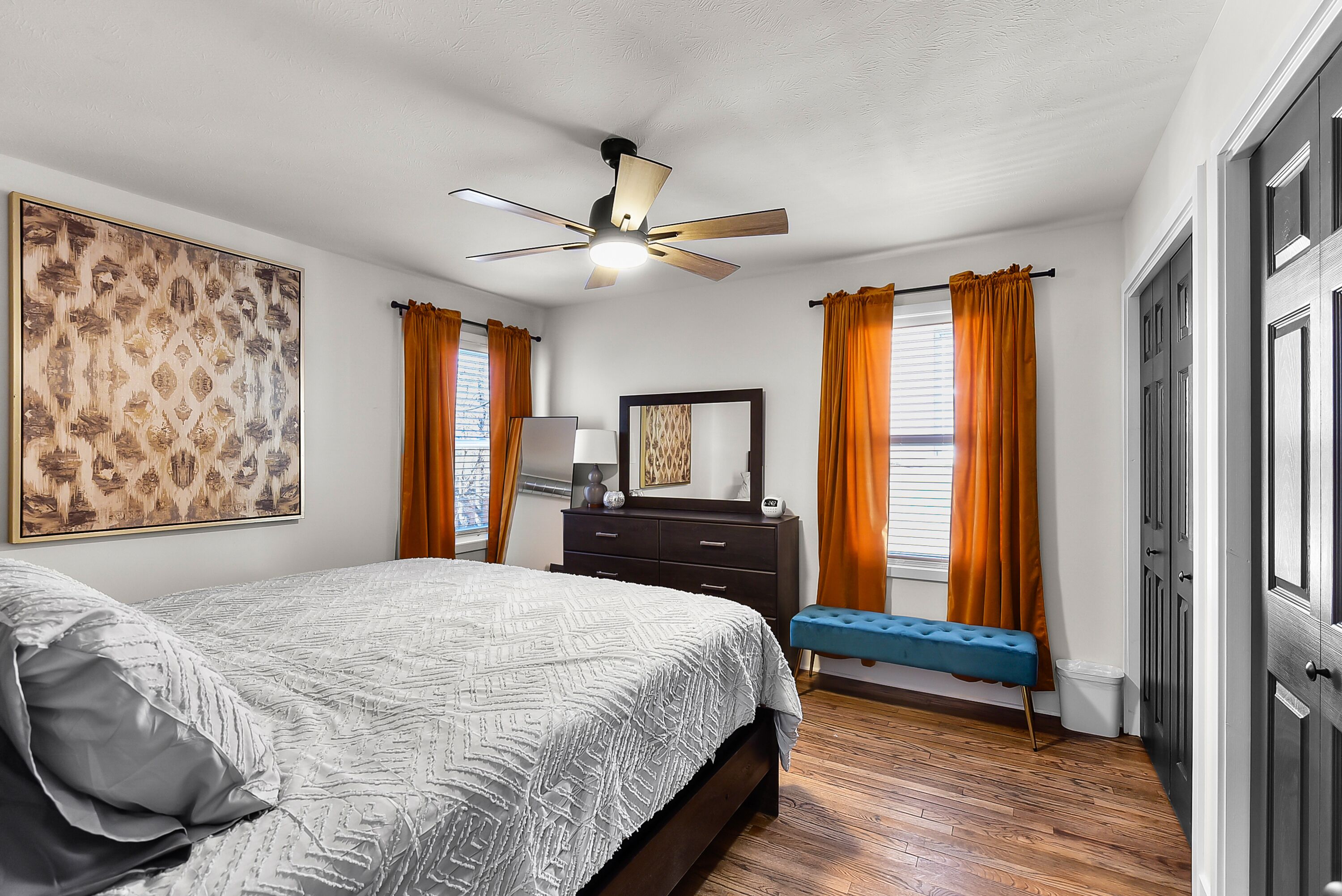 A bedroom with a bed, a dresser, a bench, a ceiling fan, and a large framed picture on the wall.