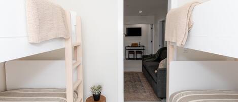 A room with bunk beds, a small stool with a plant, and a doorway leading to a living area with a sofa and a TV.
