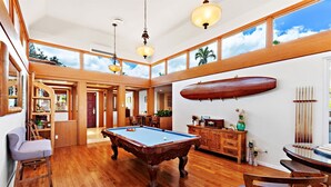 Amplia habitación con mesa de billar, muebles de madera y grandes ventanales con vistas al cielo y a los árboles.