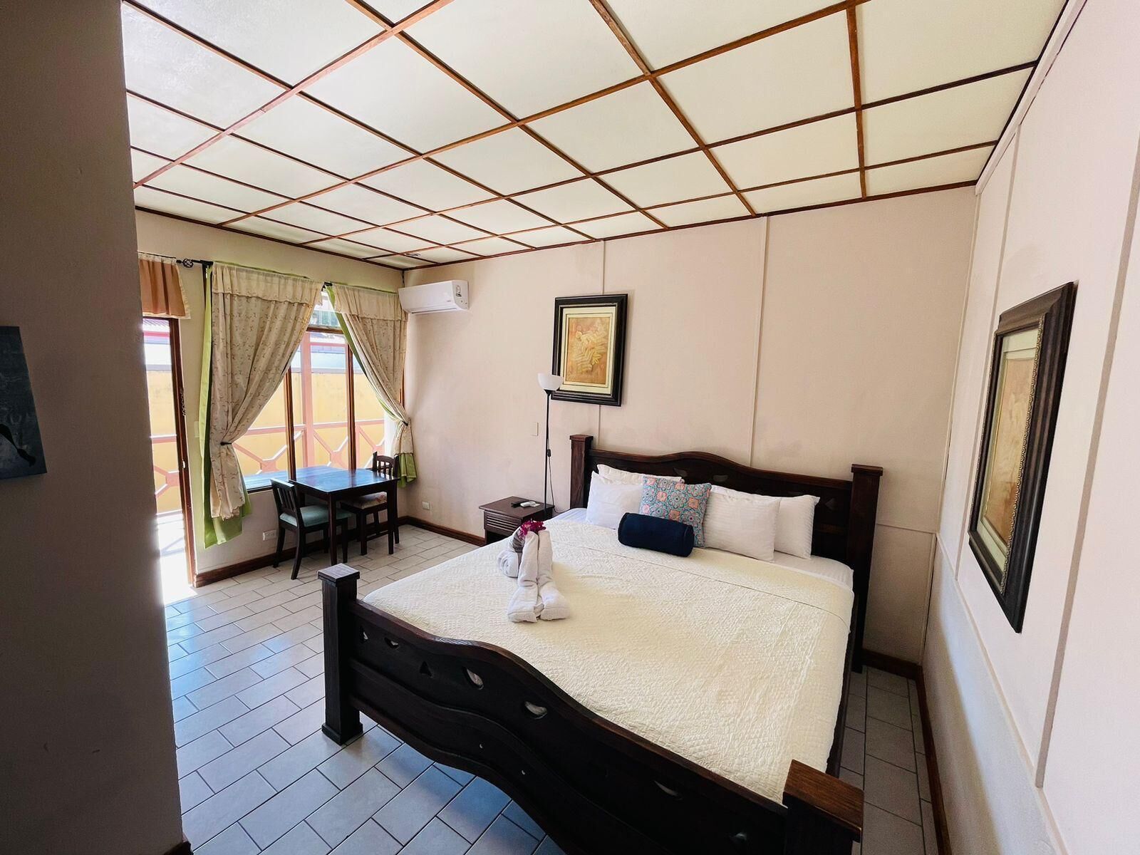 A hotel room with a bed, a desk, and a chair. There is a window with curtains, a painting on the wall, and a tiled floor.