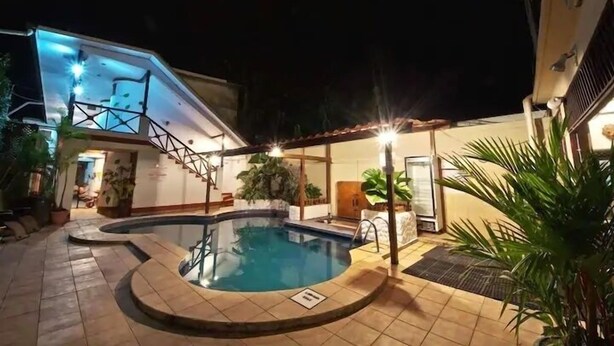A swimming pool with a tiled deck, surrounded by potted plants and a staircase leading to a balcony. - Poseidon Party Hotel In Downtown (Jacó)