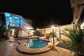 Une piscine dotée d’une terrasse carrelée, entourée de plantes en pot et accessible par un escalier menant à un balcon.