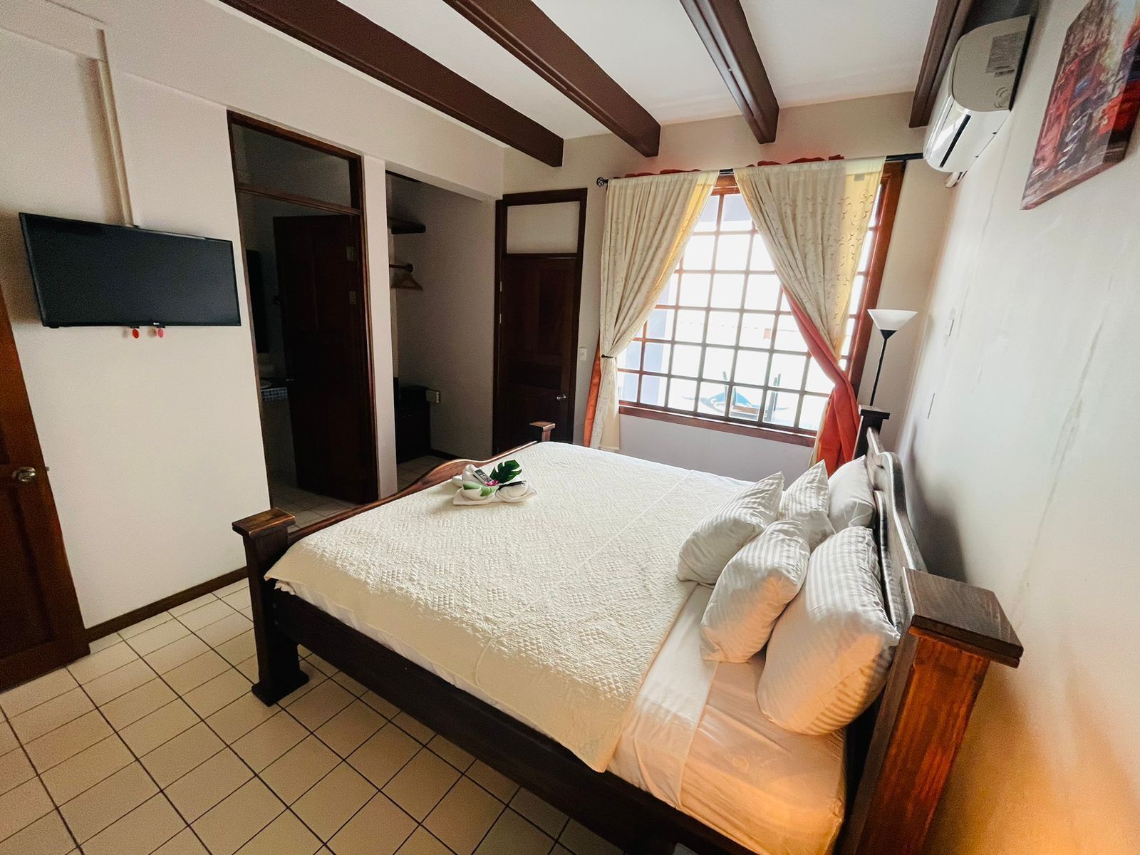 A neatly arranged bedroom with a bed, a television mounted on the wall, and a window with curtains.
