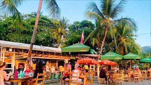 An outdoor restaurant with wooden tables and chairs, surrounded by palm trees and umbrellas. - Poseidon Party Hotel In Downtown (Jacó)