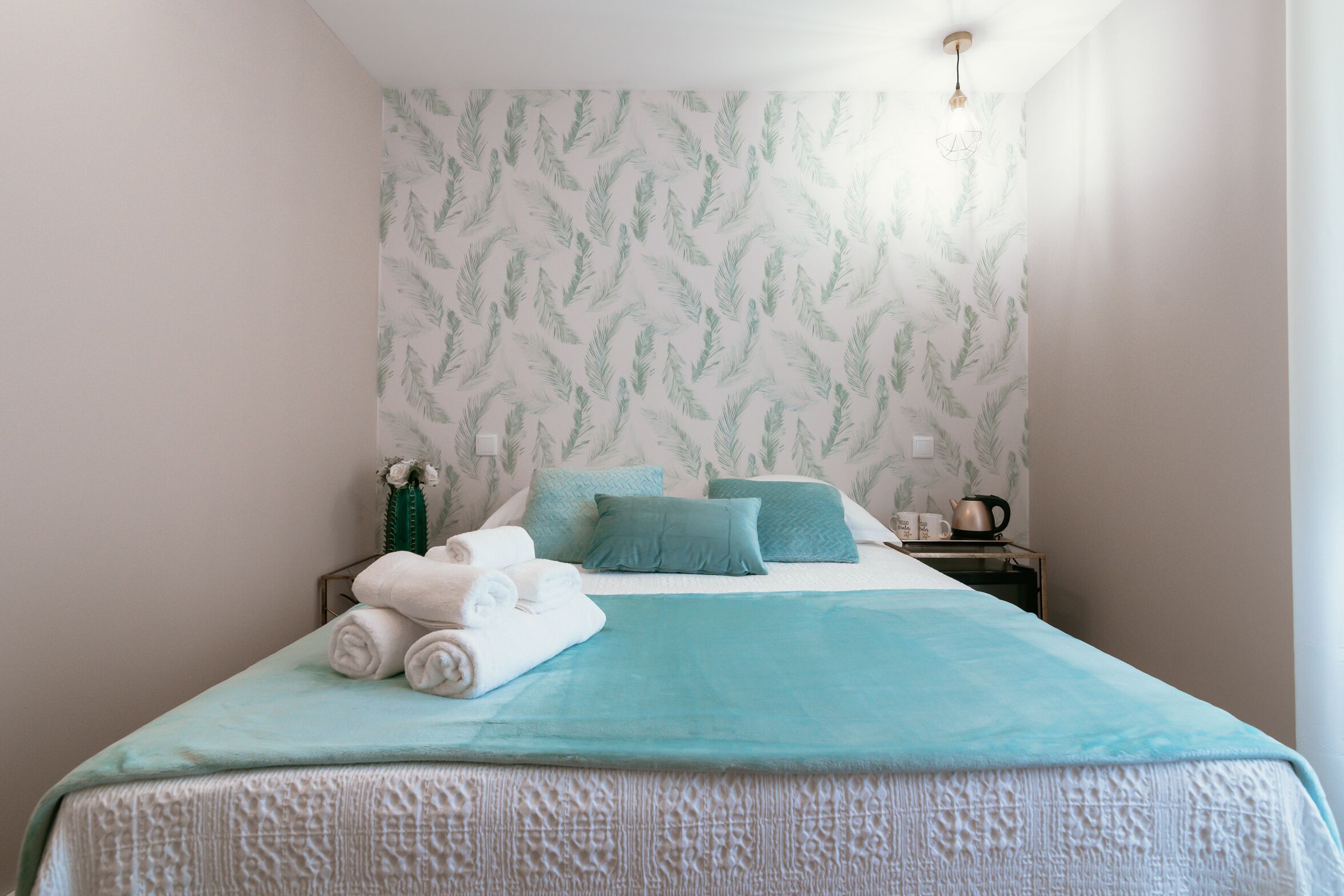 A neatly made bed with a teal bedspread and pillows, a small bedside table with a kettle, and a wall with a leafy pattern.