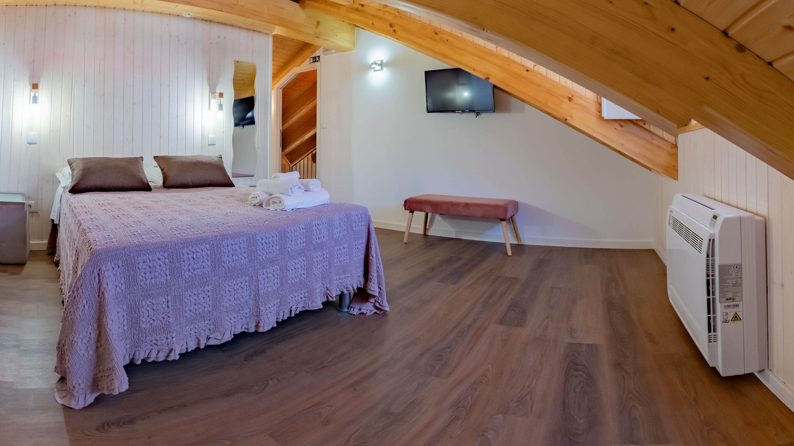 Un dormitorio con techo de madera, una cama con manta rosa, un banco, un televisor instalado en la pared y un radiador blanco.
