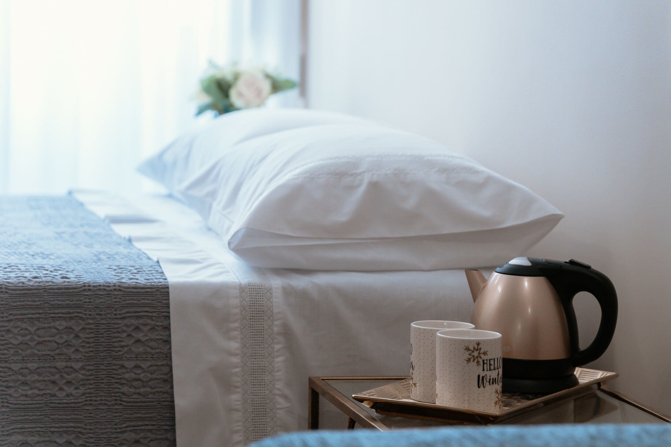 A neatly made bed with white bedding, a bedside table with a kettle and two mugs, and a window with a view of greenery.