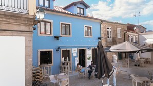 Un café en plein air avec des tables et des chaises, un bâtiment bleu avec un toit en tuiles, et une enseigne sur laquelle on peut lire « FAMAS ».
