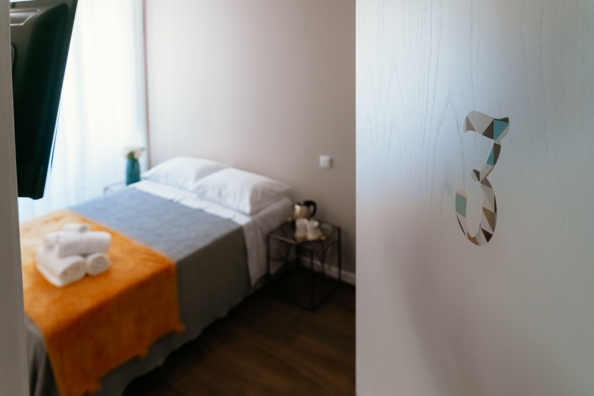 Hotellihuone, jossa on sänky, pieni pöytä, jonka päällä on koriste-esine, ja seinä, jossa on numero ’3’ -tarra.
