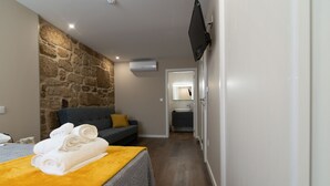 Une chambre d’hôtel avec un mur décoratif en pierre, un canapé gris agrémenté d’un coussin jaune, un lit recouvert d’une couverture jaune et un téléviseur fixé au mur.