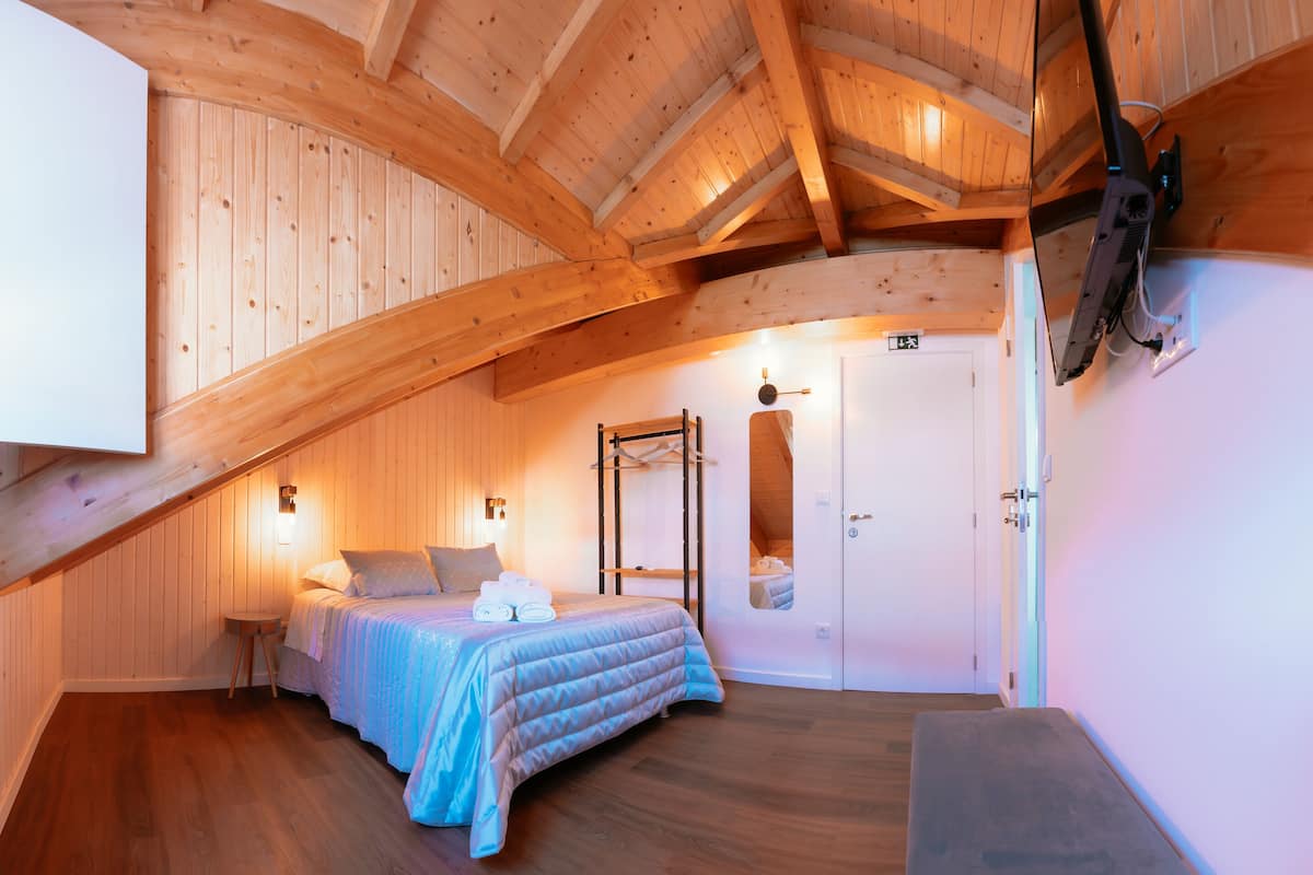 Une chambre avec un plafond en bois, un lit recouvert d’une couverture bleue, une table de chevet et un téléviseur fixé au mur.