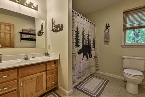 Une salle de bain avec un meuble-lavabo en bois, des toilettes, un rideau de douche orné de motifs représentant une forêt et des animaux, et une fenêtre avec des stores.