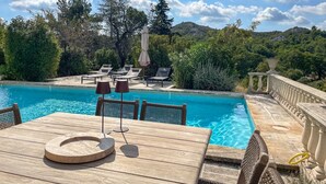Zona junto a la piscina con mesa de madera, sillones de descanso y vistas a un paisaje montañoso.