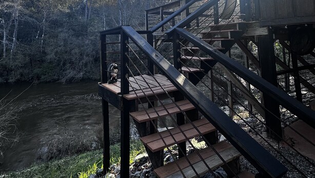 A wooden staircase leading to a covered area with an American flag, surrounded by trees and a river.