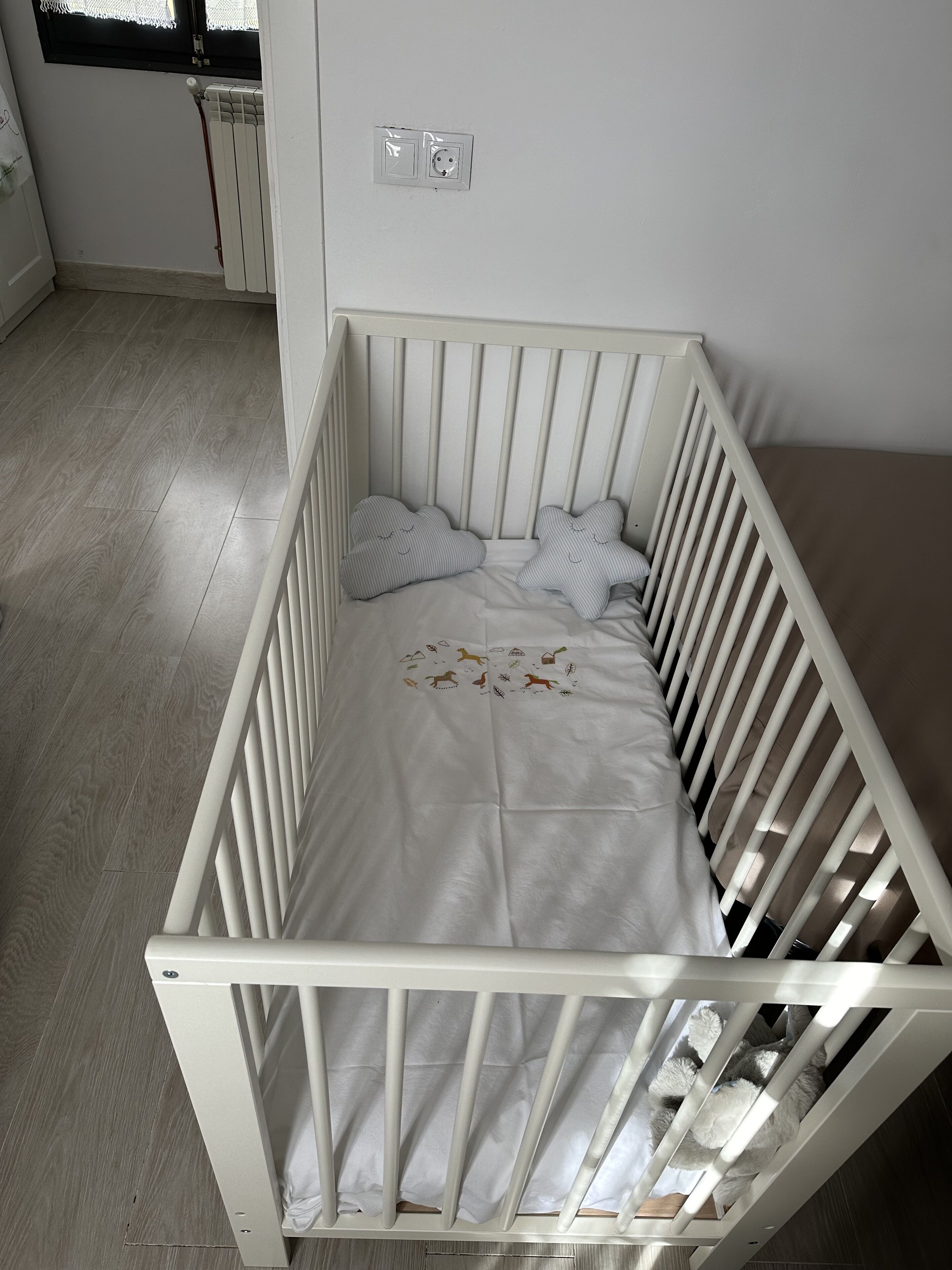 Een wit wiegje met een witte matras en gestreepte kussens, geplaatst tegen een muur met een raam erboven.