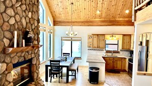 A spacious kitchen with wooden cabinets, a stainless steel refrigerator, and a dining area with a stone wall and a fireplace. - Cozy Cabin Escape: Nature, Comfort, and Serenity Await! (Grand Lake)