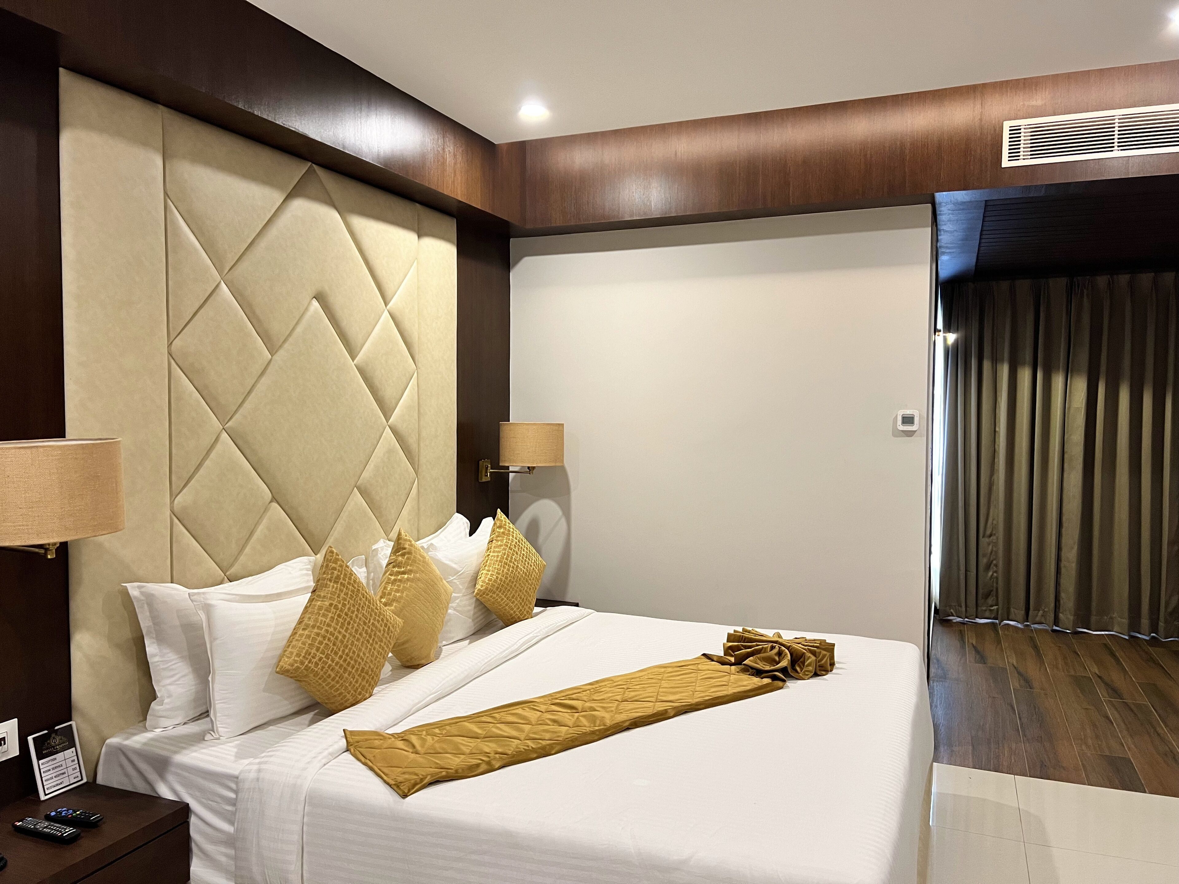 A hotel room with a large bed, a headboard with a diamond pattern, a bedside lamp, and a nightstand with a remote control.