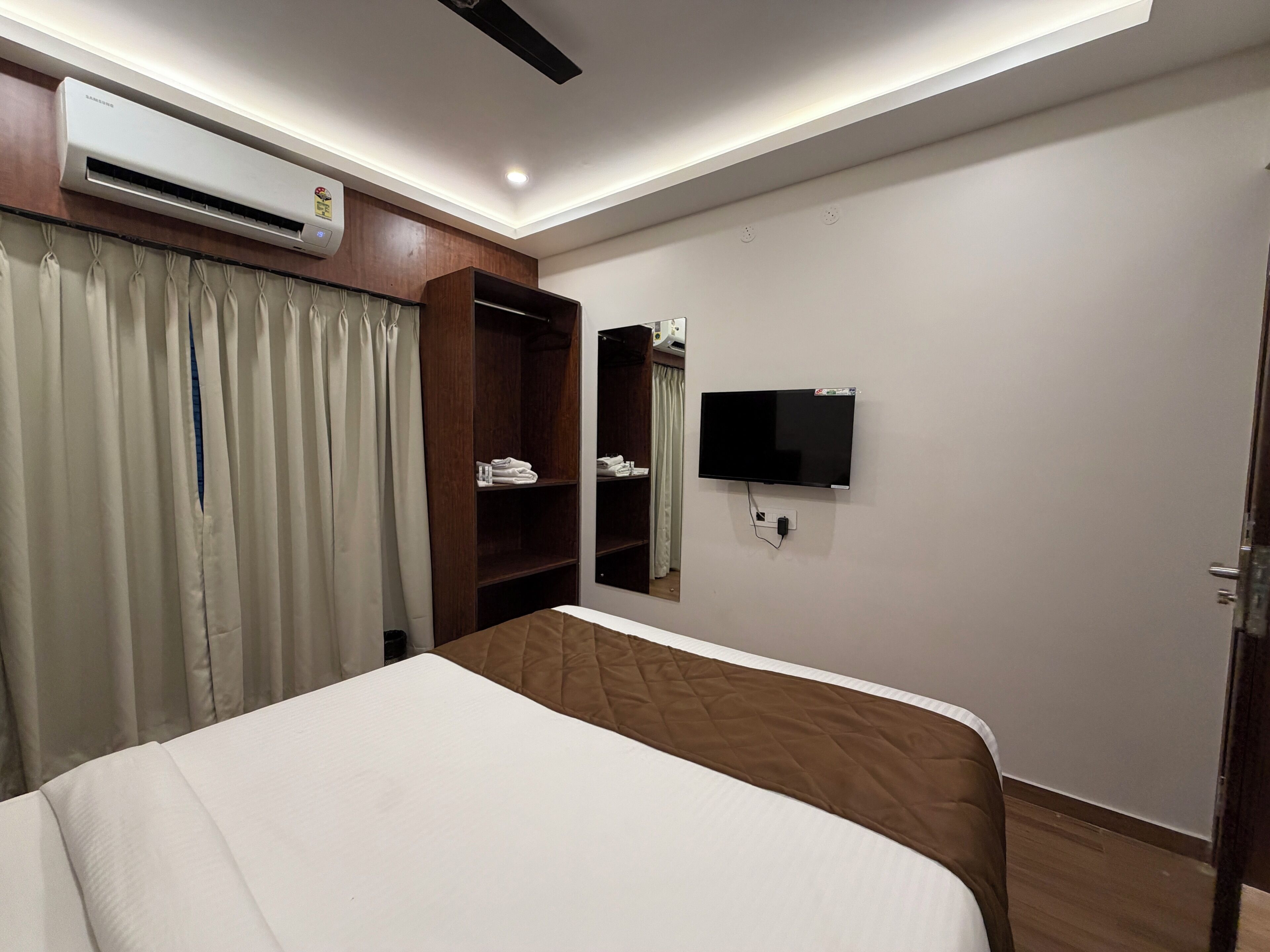A hotel room with a bed, a TV mounted on the wall, a wardrobe, and an air conditioning unit.