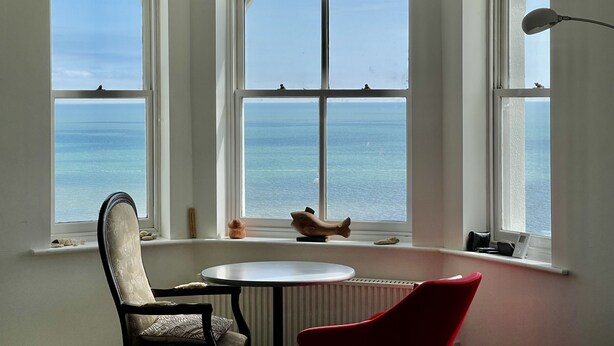 Ein Zimmer mit Erkerfenster und Meerblick, ein runder Tisch, ein roter Sessel und eine Lampe.