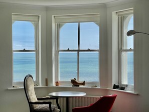 A room with a bay window overlooking the sea, a round table, a red chair, and a lamp. - Sea View Apartment, Folkestone (Folkestone)
