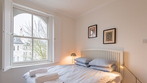 Ein Schlafzimmer mit großem Fenster, einem Bett mit weißer Bettwäsche, zwei gerahmten Bildern an der Wand und einem Nachttisch mit Lampe.
