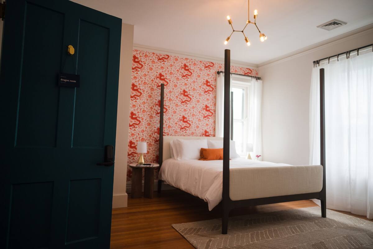 A bedroom with a four-poster bed, a nightstand with a lamp, a window with curtains, and a teal door.