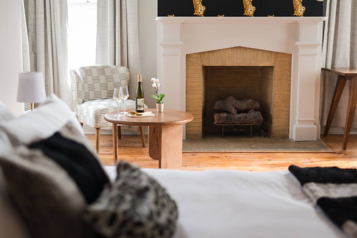 A hotel room with a fireplace, a round wooden table with a bottle of wine and glasses, a chair, and a bed with a black and white throw blanket.