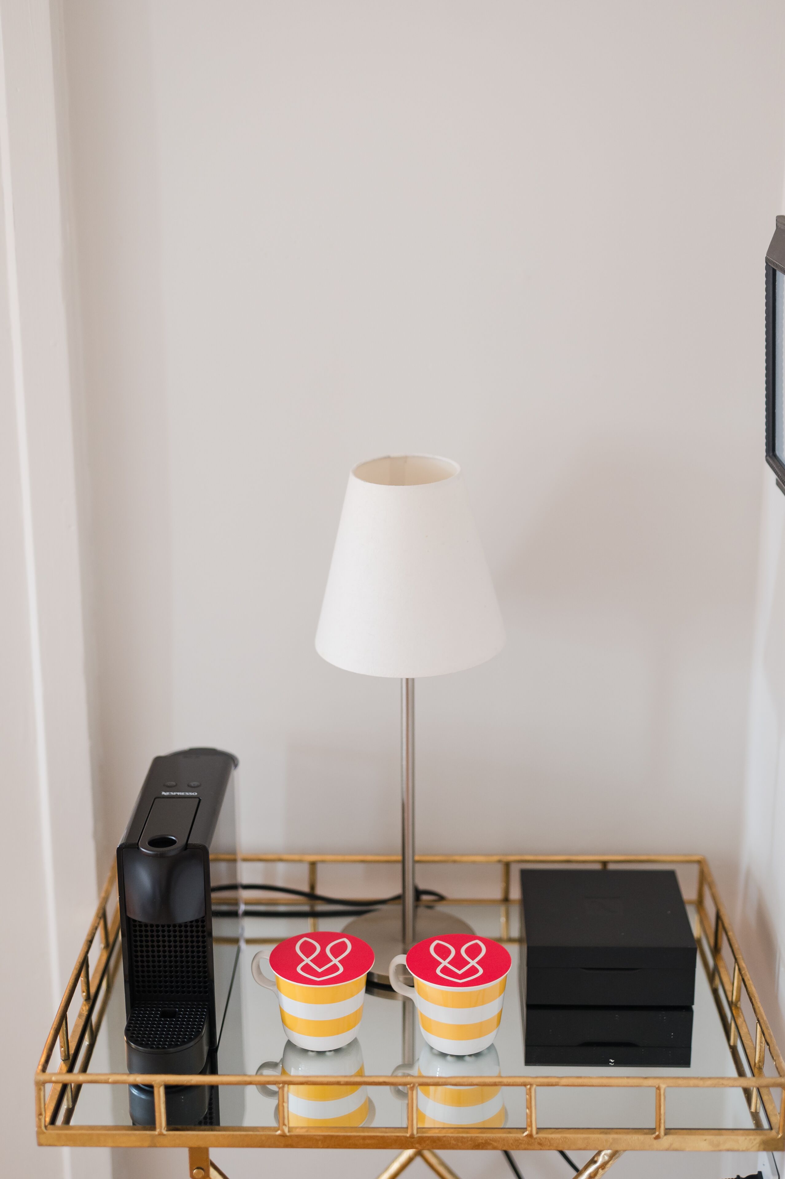 Een tafeltje met een lamp, een zwart elektronisch apparaat, twee gestreepte mokken en een zwarte doos.