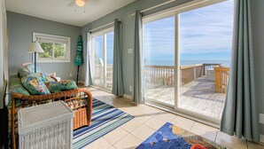 A room with a large sliding glass door leading to a wooden deck with a view of the ocean. There is a wicker sofa with colorful cushions and a small table with a lamp. - Virginia's Beach - Cozy Oceanfront Beach Cottage! (Surf City)