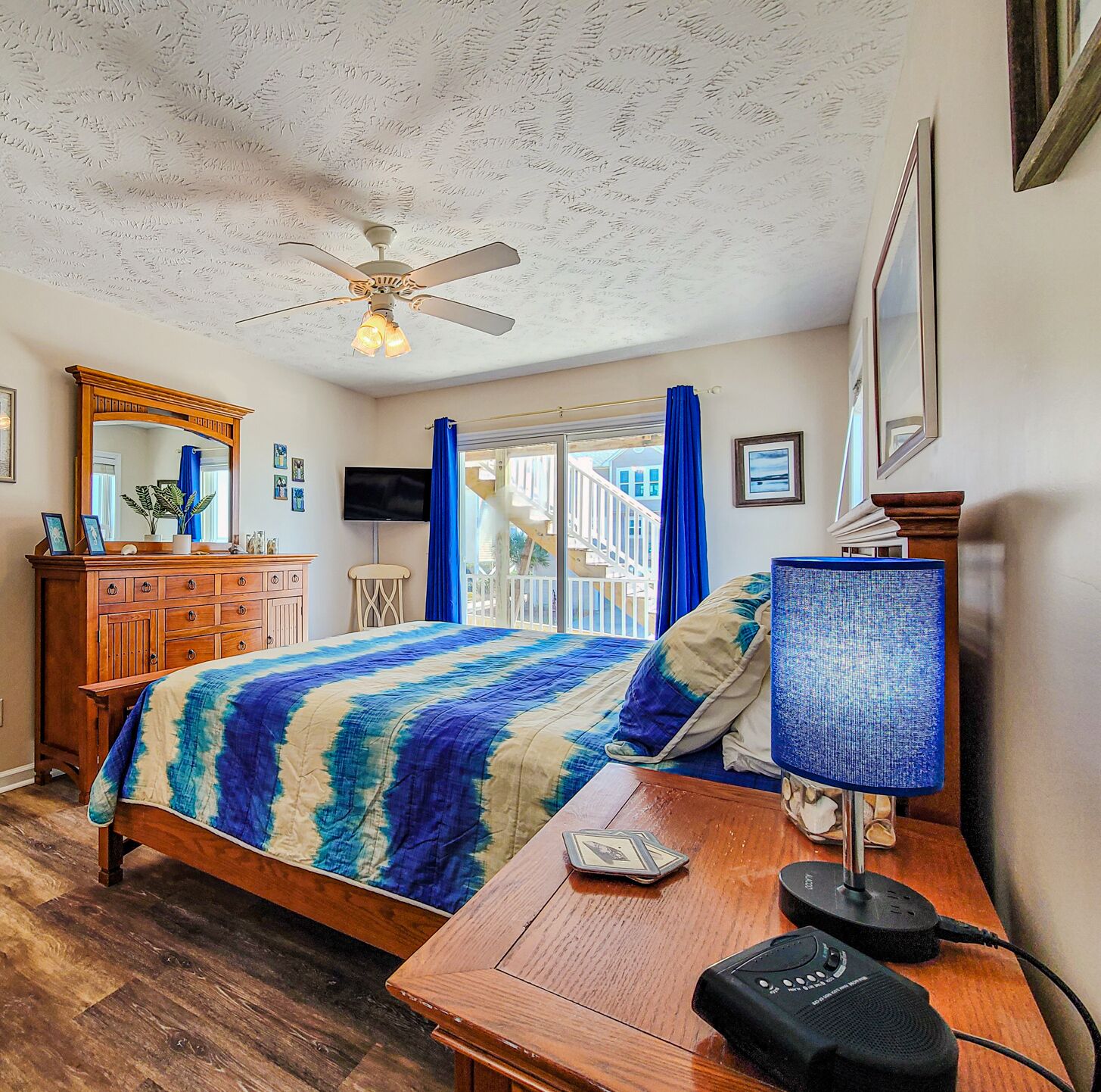 A bedroom with a bed, a wooden dresser, a ceiling fan, a TV, and a window with curtains.