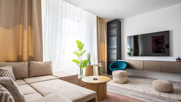 Ett modernt vardagsrum med en soffa, ett runt soffbord, en blå fåtölj, en tv och ett stort fönster med gardiner.