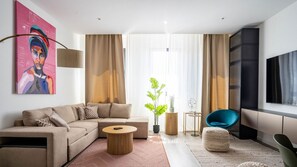 A modern living room with a beige sofa, a round coffee table, and a blue chair. There is a large window with sheer curtains, a potted plant, and a painting on the wall.