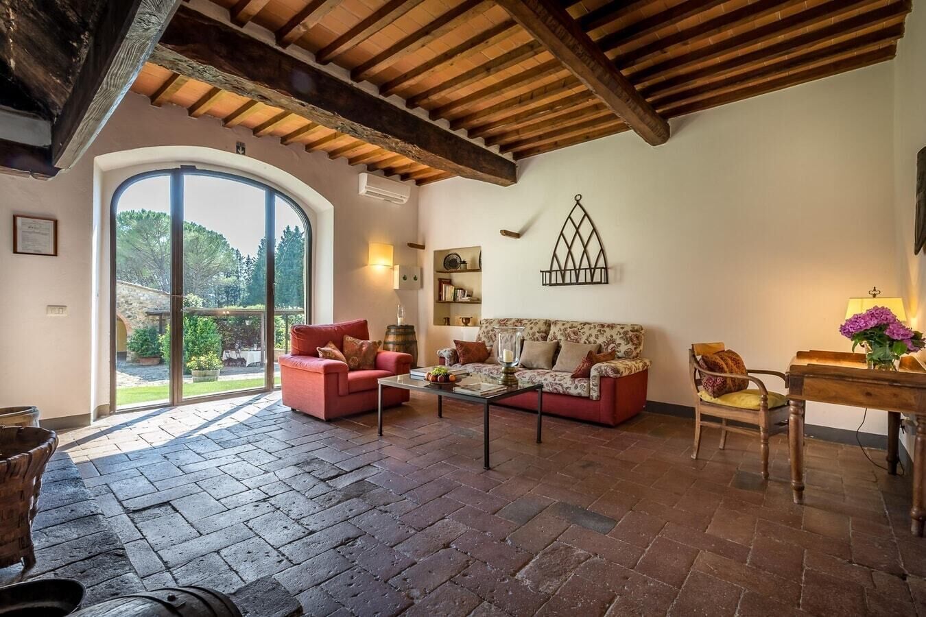 A spacious living area with a red sofa, a patterned sofa, and a wooden chair. There is a coffee table with decorative items, a fireplace, and a large window with a view of greenery.