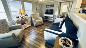 A living room with a ceiling fan, a TV, a sofa, a chair, and a table with decorative items. - The Nautical Nook Beach Cottage - #1714 Ocean Lakes (Myrtle Beach)