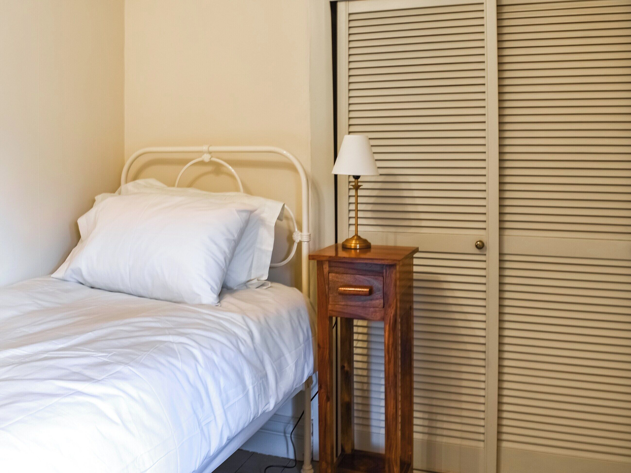 A single bed with white bedding, a bedside table with a lamp, and a wooden drawer.
