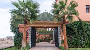 A gated entrance with a tiled roof, palm trees, and a brick pathway leading to a building with a pink facade. - Yanis Guest House  (Marrakech)