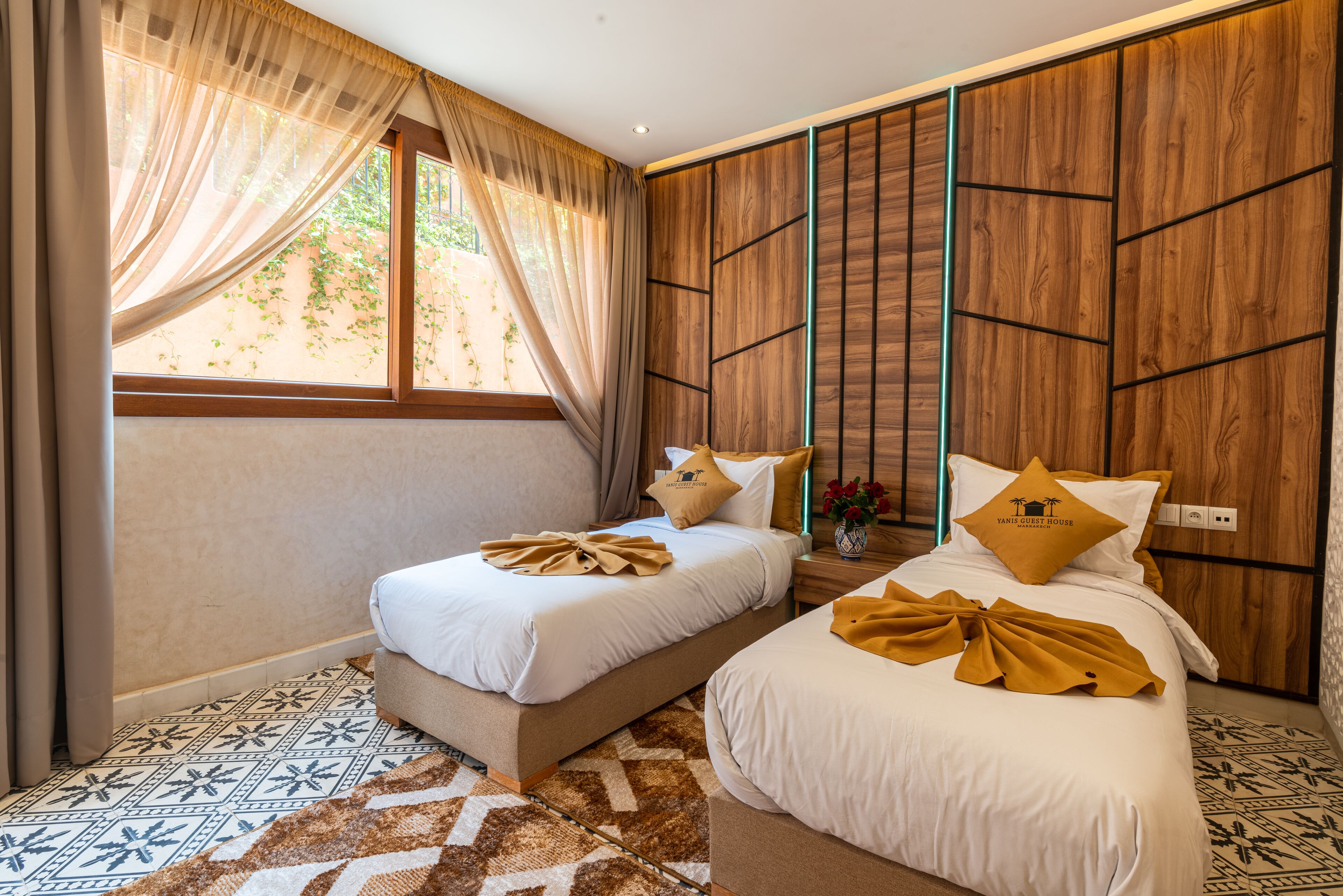 A hotel room with two beds, a large window with curtains, a patterned floor, and wooden paneling on the walls.
