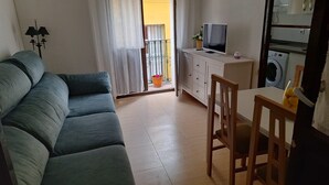 A living room with a sofa, a TV on a cabinet, a washing machine, and a balcony with a view. - Feria Especial (Sevilla)