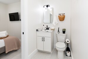 A modern bathroom with a white sink, mirror, and toilet. There is a basket on the wall and a small plant on the toilet. - The Inn at Fulton (Sandusky)